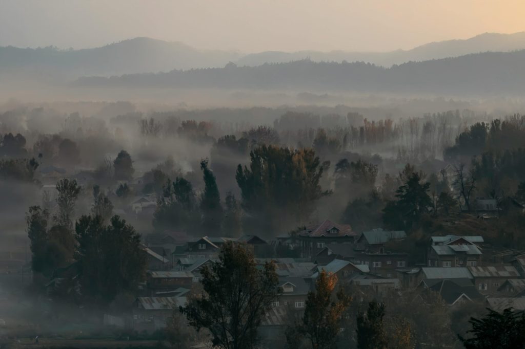 A serene, mist-filled morning scene in Srinagar with village rooftops and distant mountains.